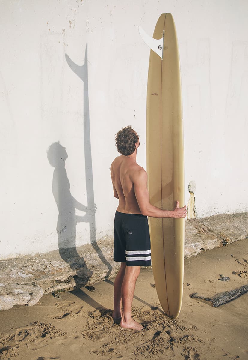 Man holding a surfboard on a sandy beach with a white wall in the background