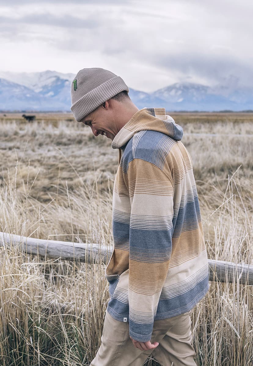 Man wearing a plaid shirt and beanie standing in a field with mountains in the background